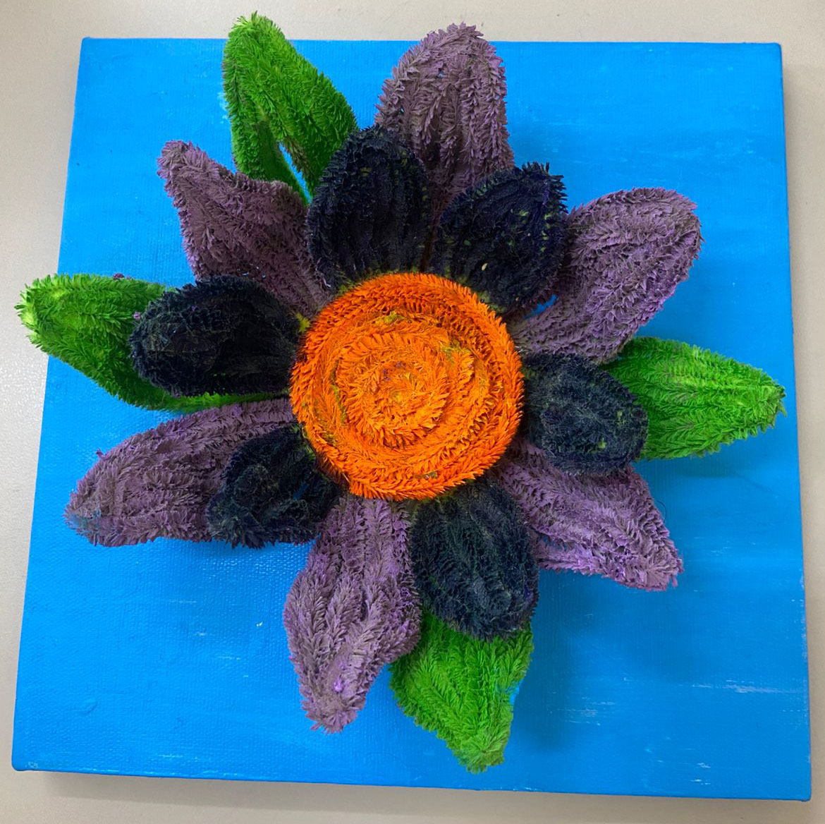 Flor com pétalas verdes, roxo-claro e roxo-escuro feita com limpadores de cachimbo, sobre fundo azul pintado.