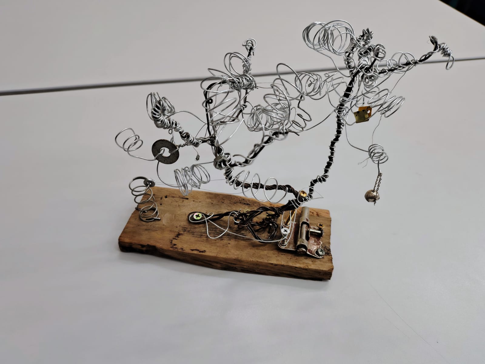 Escultura abstrata feita de fios metálicos retorcidos, com peças adicionais como parafusos e molas, fixada em uma base de madeira rústica. A forma lembra um galho ou árvore