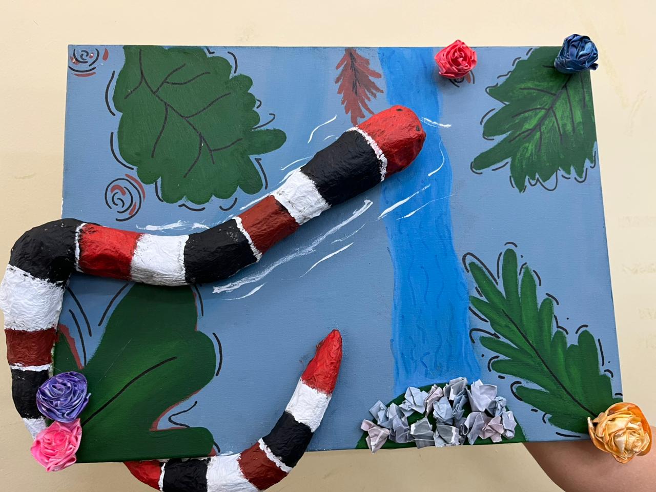 Obra com textura vibrante: rio azul central, folhas verdes, cobra tridimensional colorida, flores e papéis que dão relevo e naturalidade