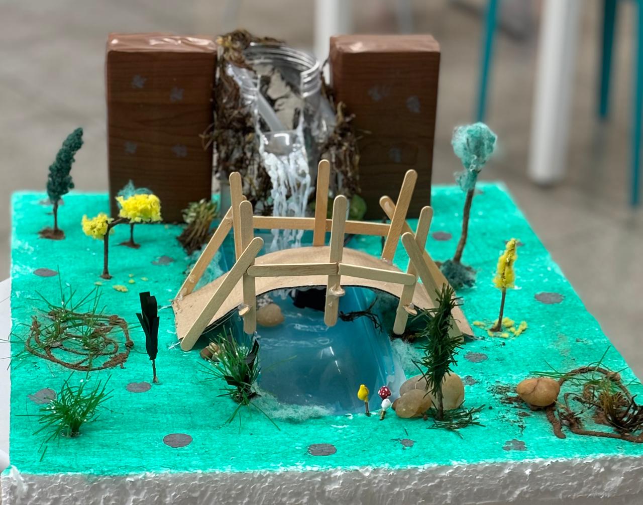  Maquete com ponte de palitos sobre rio artificial, cachoeira de algodão, árvores feitas à mão e gramado verde em base de isopor