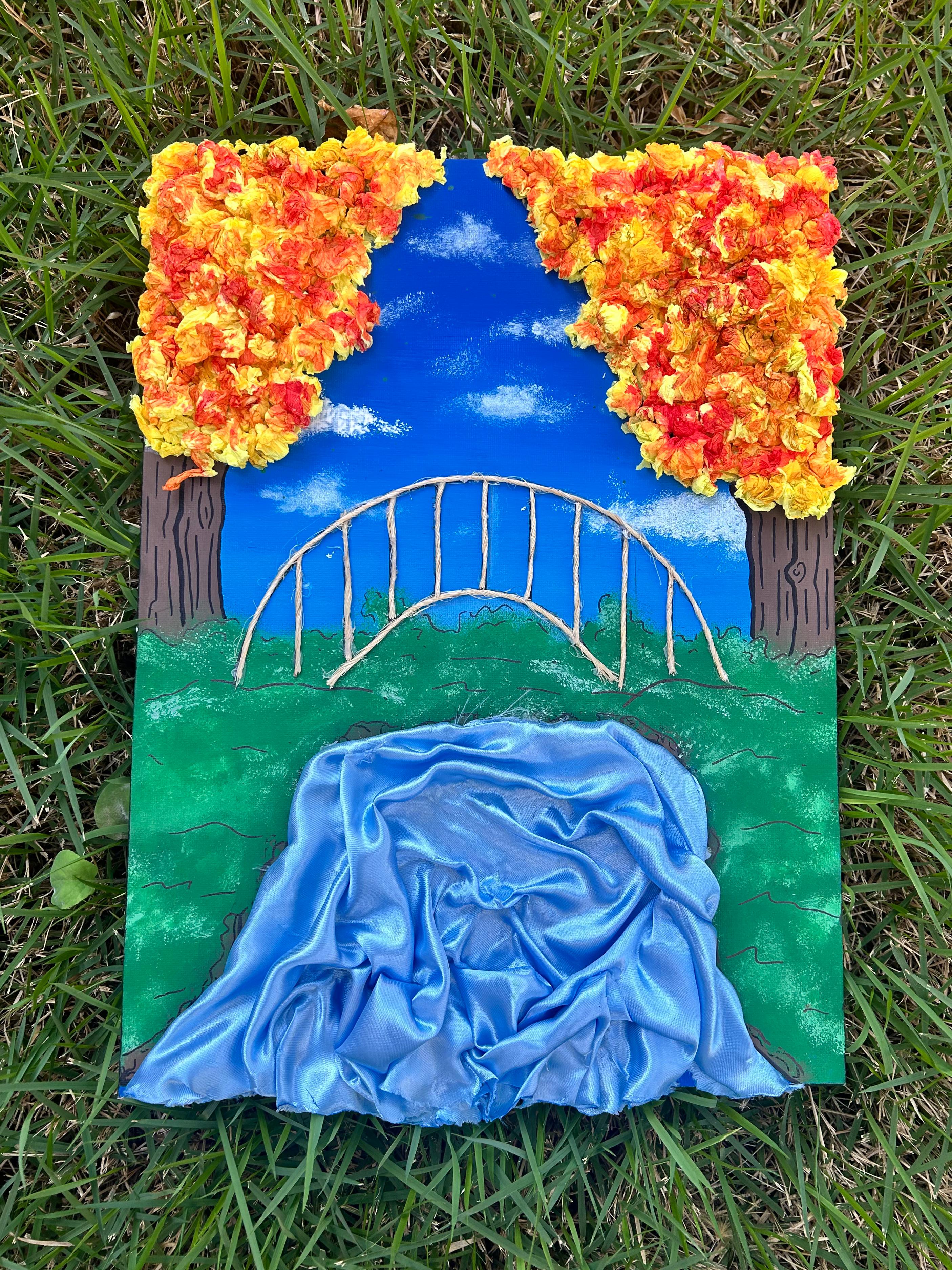 Pintura de outono com céu azul, árvores de papel crepom em tons de vermelho, amarelo e laranja, e uma ponte arqueada de corda sobre um rio de cetim.