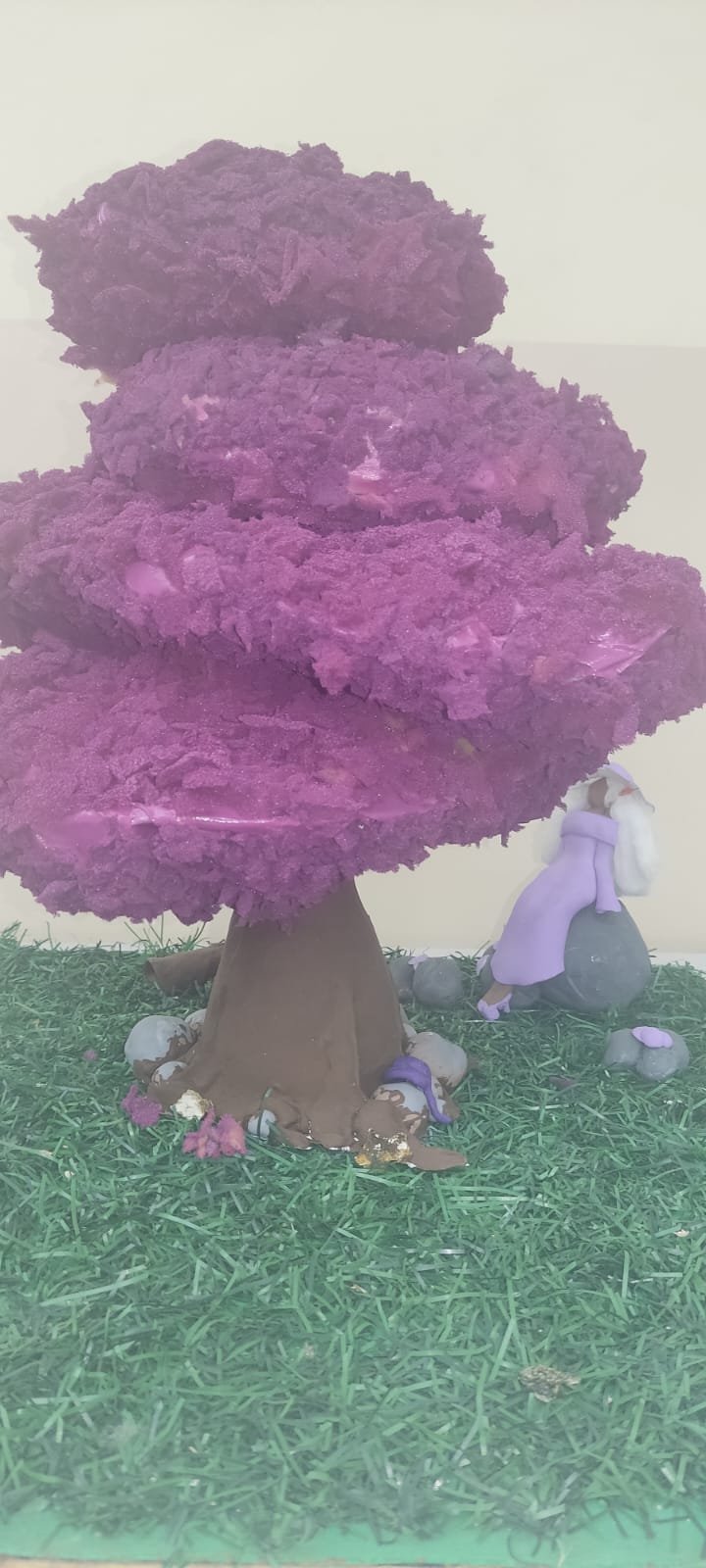 Escultura tridimensional de uma mulher negra de cabelos brancos sentada sobre uma pedra cinza com cogumelos violetas, ao lado de uma árvore de tronco marrom e folhagem roxa